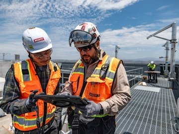 Two people wearing construction PPE at a jobsite while looking at a tablet. 